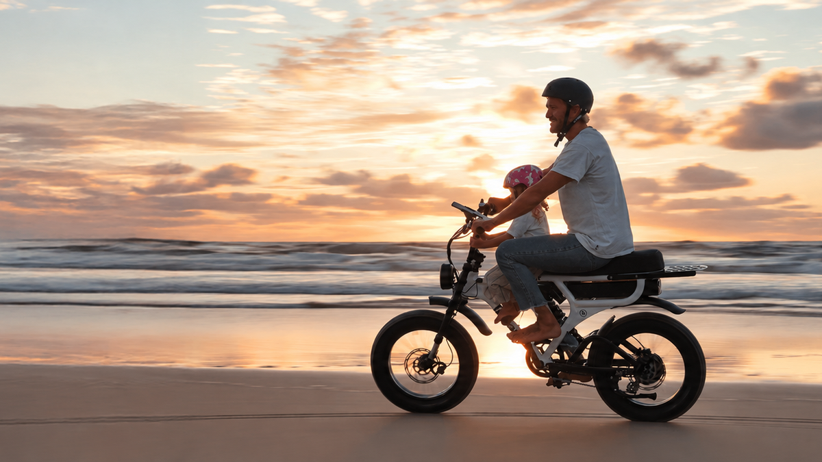 A man rides an Fatboy electric bicycle along a sandy beach in Shellharbour at sunset, with ocean waves and a colorful sky in the background.